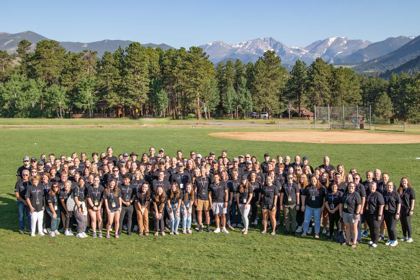 Grupo grande afuera en un campo de béisbol con montañas al fondo